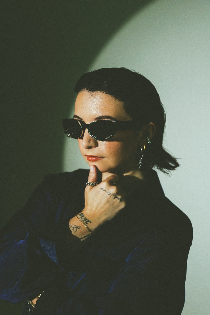 women with slick back hairstyle poses with hand on chin with sunglaases on wearing black turtle neck long sleeve on grey backdrop